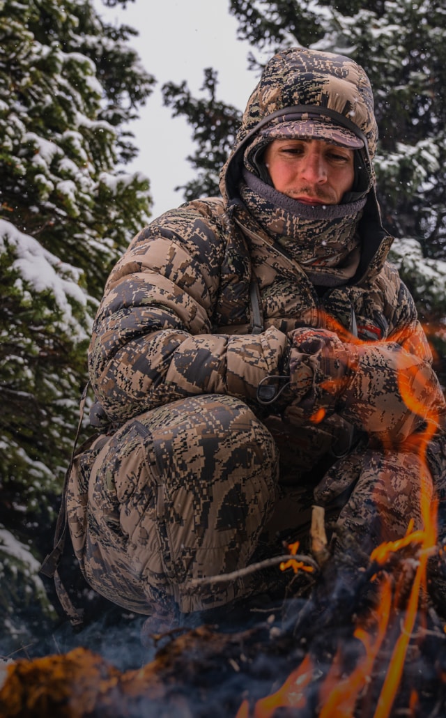 Adam Foss sitting by a camp fire in the HyperDown Jacket in Optifade Open Country | SITKA Gear Holiday Gift Guide